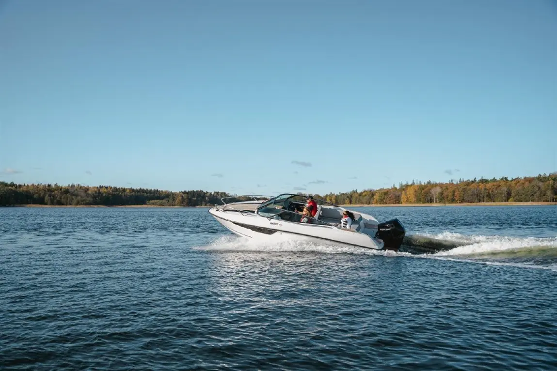 A white speedboat with four people is skimming across a calm blue lake on a sunny day with trees in the background.